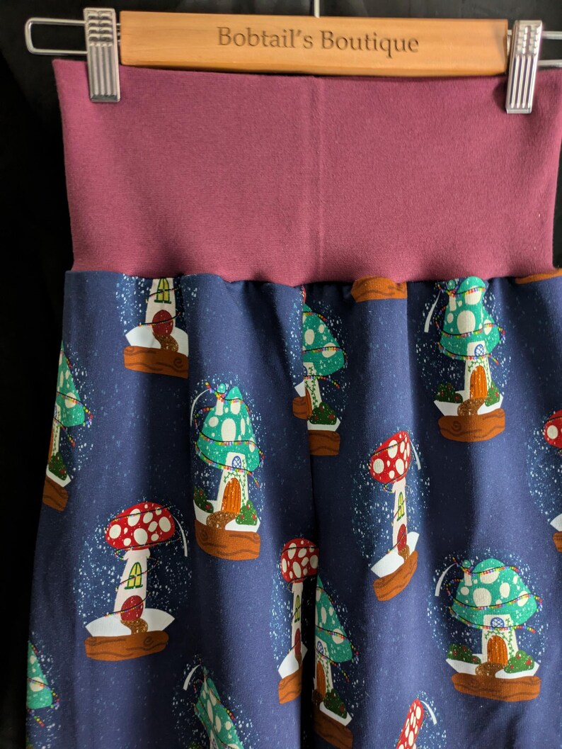 May include: Blue pants with a repeating pattern of mushroom houses in red, green, and white. The waistband is a solid mauve color. The pants are hanging on a wooden hanger with the text "Bobtail's Boutique".