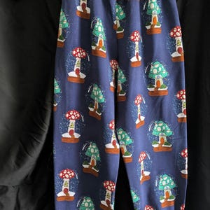 May include: Blue pants with a repeating pattern of mushroom snow globes. The pants have a wide, mauve waistband and matching cuffs. The text "Bobtail's Boutique" is visible on the hanger.