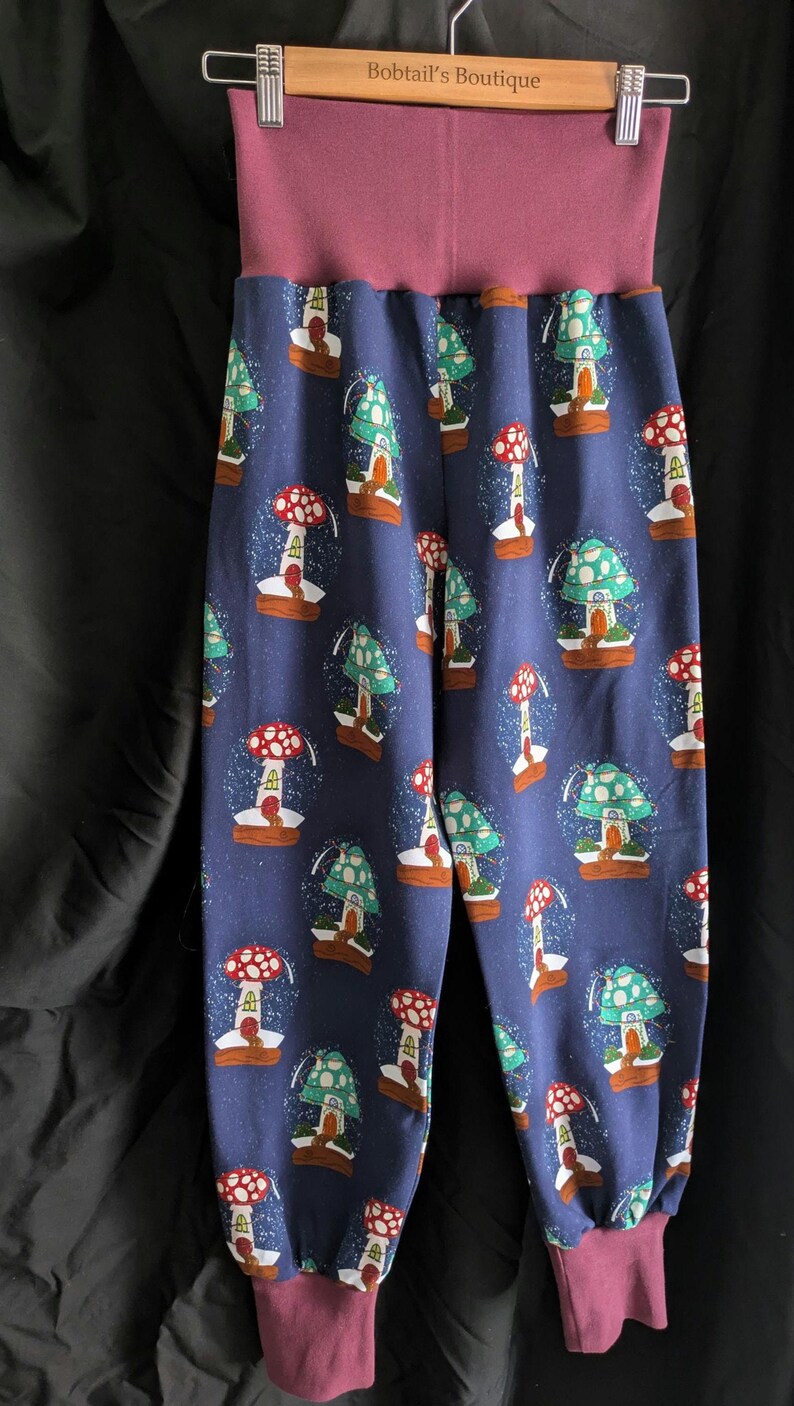 May include: Blue pants with a repeating pattern of mushroom snow globes. The pants have a wide, mauve waistband and matching cuffs. The text "Bobtail's Boutique" is visible on the hanger.