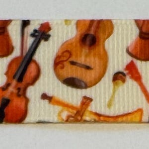 May include: Cream-colored ribbon featuring a repeating pattern of musical instruments. The design includes violins, guitars, saxophones, maracas, and drums in shades of brown and orange.