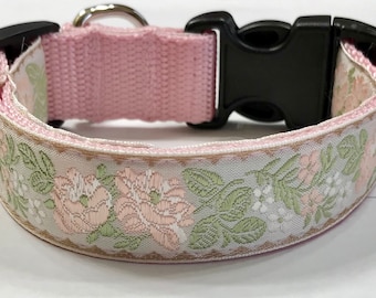 May include: A pink and white dog collar with a floral pattern. The collar has a black plastic buckle.