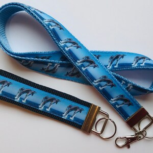 May include: A blue lanyard and wristlet featuring a repeating pattern of leaping dolphins against a blue and white striped background. The wristlet has a black base and silver hardware. The lanyard has a silver clasp.