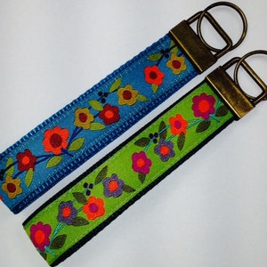 May include: Two keychains with floral patterns. One keychain has a blue background with red, orange, and yellow flowers. The other keychain has a green background with orange, pink, and purple flowers. Both keychains have brass key rings.