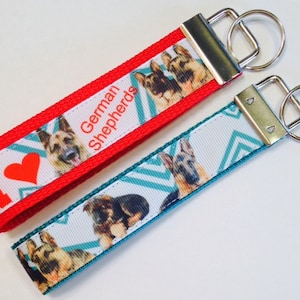 May include: Two keychains with a red and white ribbon with the text "I love German Shepherds" and a teal and white ribbon with a geometric pattern. Both keychains feature images of German Shepherds.