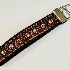 May include: A decorative key fob with a dark red background and a floral pattern in white and green. The key fob has a silver metal clasp and a key ring.