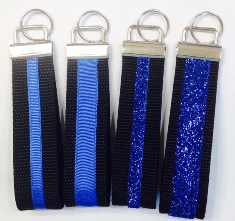 May include: Four keychains with black fabric and a blue stripe. The stripes are different styles: a solid blue ribbon, a blue ribbon with a textured weave, a blue glitter stripe, and a blue glitter stripe with a textured weave.