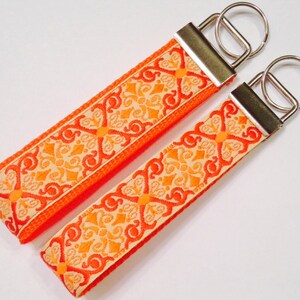 May include: Two orange and cream patterned keychains with silver key rings. The keychains are made of fabric with a repeating geometric design.
