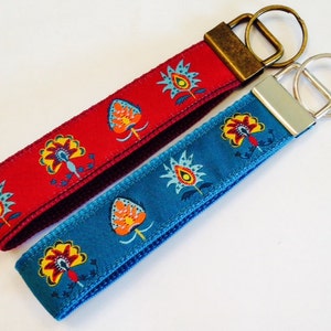 May include: Two keychains with decorative floral patterns. One keychain is red with a bronze-colored ring, and the other is teal with a silver-colored ring. The floral pattern includes yellow, orange, and blue details.