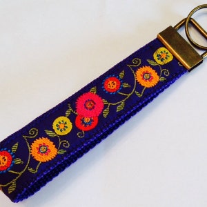 May include: A dark blue key fob with a floral design. The key fob features embroidered flowers in shades of pink, yellow, and orange, with green leaves and stems. The key fob has a bronze-colored metal clasp and key ring.