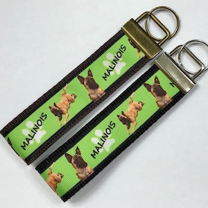May include: Two green and black keychains with a brown dog breed, the Malinois, printed on them. The keychains have the word "Malinois" printed on them with a paw print.