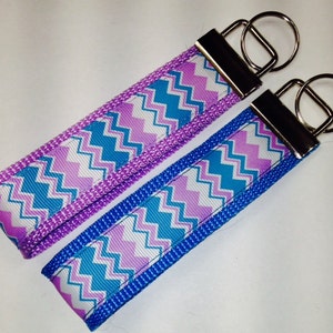 May include: Two keychains with a chevron pattern in shades of purple, blue, and white. The keychains are made of fabric and have silver key rings.