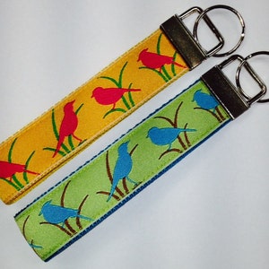 May include: Two keychains with colorful bird designs. One keychain has a yellow background with red birds and green grass. The other keychain has a green background with blue birds and brown grass.