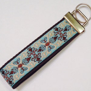 May include: A brown key chain with a blue and gold patterned ribbon. The ribbon has a repeating pattern of brown, blue, and gold swirls. The key chain has a gold metal ring.