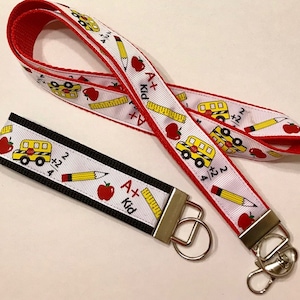 May include: A red and white lanyard with a school bus, pencil, apple, and ruler design. The lanyard has a black and white key chain with the same design. The text on the lanyard reads "A+ Kid".