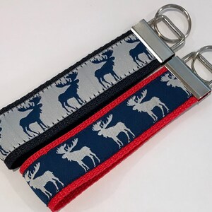 May include: Two keychains with silver key rings. One keychain has a black strap with a silver moose pattern on a gray background. The other keychain has a red strap with a navy blue moose pattern on a silver background.