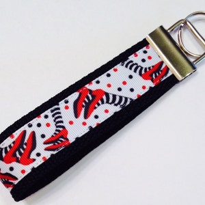 Puede incluir: Un llavero blanco y negro con un patrón de zapatos de bruja rojo y negro. El llavero tiene un llavero plateado.