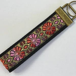 May include: A black wristlet key fob with a floral design in shades of red, pink, and gold. The key fob has a bronze-colored metal ring and clasp for attaching keys. The floral pattern is woven into the fabric.