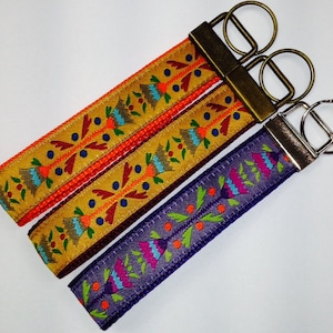 May include: Three colorful keychains with intricate patterns and metal key rings. The keychains are made of woven fabric with a variety of colors, including orange, yellow, purple, green, and blue.