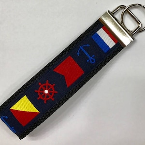May include: Navy blue keychain strap with embroidered nautical flags, a ship's wheel, and an anchor. The strap has a black border and a silver metal key ring.