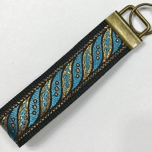 May include: A black key chain with a teal and gold patterned ribbon. The key chain has a brass key ring.