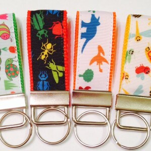 May include: Four colorful keychains with different designs. Each keychain features a white background with a colorful pattern of insects, dinosaurs, or caterpillars. The keychains are attached to silver key rings.