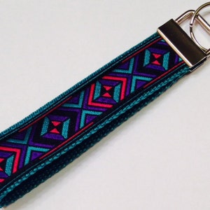 May include: A teal key fob with a geometric pattern in purple, red, and teal. The key fob has a silver metal clasp and a metal ring for keys. The design is a repeating pattern of squares and crosses.