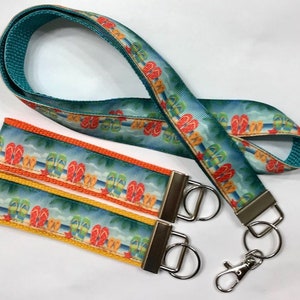 May include: A teal blue lanyard with a colorful print of flip flops on a beach. The lanyard has a matching key chain with two silver key rings and a silver lobster clasp.