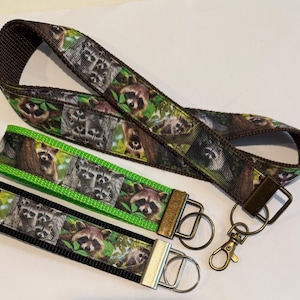 May include: Three raccoon-themed wristlet keychains in brown, green, and black. The keychains feature a repeating pattern of raccoons in a forest setting. Each keychain has a metal clasp and ring for attaching keys.