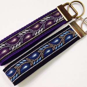 May include: Two keychains with patterned fabric straps and metal key rings. One strap is purple with silver and gold accents, the other is black with blue and silver accents.
