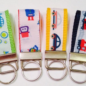 May include: Four colorful keychains with different designs. Each keychain features a white background with a repeating pattern of a different vehicle. The keychains are attached to silver key rings. The designs include a blue airplane, a red robot, a blue car, and a red train.