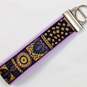 May include: A purple key chain with a silver key ring. The key chain has a black and gold patterned fabric with geometric designs.
