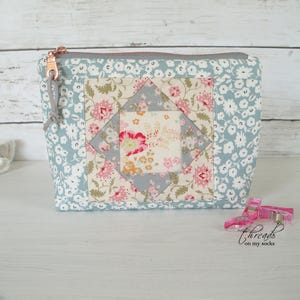 May include: A rectangular fabric zipper pouch featuring a light blue floral print background and a central square patchwork design with pink, cream, and grey floral patterns. The pouch has a copper-colored metal zipper with a grey fabric pull tab.