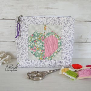 May include: A zippered pouch featuring a white fabric with a grey floral print and a central square block displaying a pieced apple design in green, pink, and white patterns. A purple cord is attached to the zipper pull, and the text threads on my socks is visible.