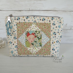 May include: A zippered pouch featuring a cream-colored fabric with a small blue floral print. The center panel displays a diamond-shaped patchwork design with green, pink, and floral patterns surrounding a central illustration of a person's face.