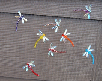 May include: A collection of nine iridescent dragonfly ornaments with beaded wings and tails in various colors, including blue, purple, orange, yellow, and red. The ornaments are displayed on a brown wooden surface.