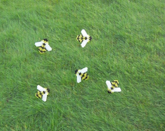 May include: Five small, decorative bee-shaped objects with white wings and yellow and black bodies are scattered across a green, grassy background. The bees appear to be made of a textured material, possibly fabric or beads.