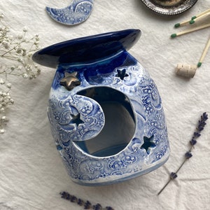 May include: Blue and white ceramic oil burner with a crescent moon design and stars. The burner has a blue glaze and a white background. It is decorated with a crescent moon and stars.