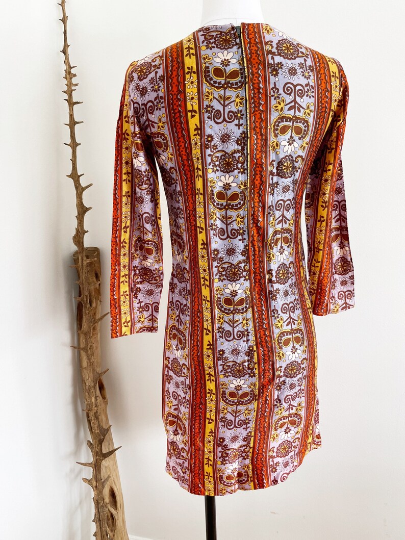 May include: A vintage 1960s dress with a colorful floral and geometric pattern. The dress has a long sleeve and a fitted silhouette. The fabric is a blend of polyester and cotton.