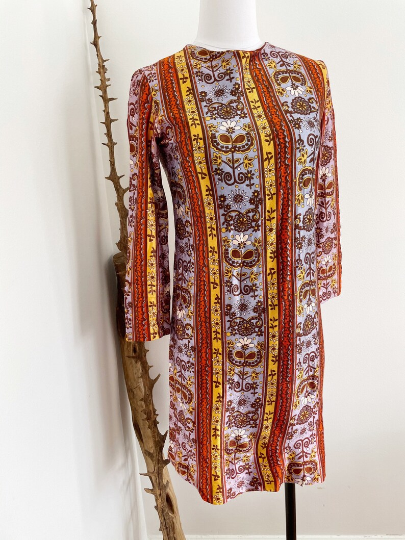 May include: A vintage 1960s style dress with a colorful floral and stripe pattern in shades of orange, yellow, brown, and purple. The dress has a long sleeve and a fitted silhouette.