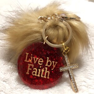 Puede incluir: Llavero de purpurina roja con un dije dorado que dice "Live by Faith" y un dije de cruz dorada con pedrería. El llavero está unido a un pompón marrón esponjoso.