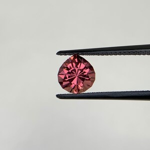 May include: A deep red gemstone with a faceted cut, held by tweezers against a white background.