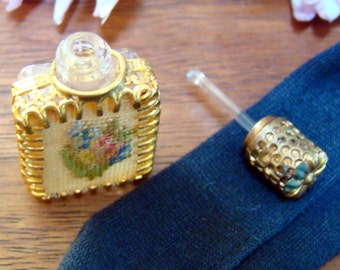 Op de afbeelding: Een klein, goudkleurig parfumflesje met een bloemenmotief en een transparante glazen bovenkant. Het flesje heeft een gouden druppelaar met een decoratieve bovenkant.