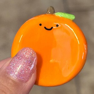 May include: A glossy orange-colored charm shaped like an orange with a smiling face. The charm has a small green leaf and a brown stem. The charm is being held by a person with a pink glitter nail.