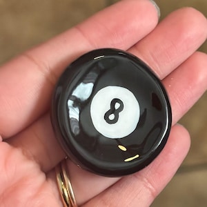 May include: A black, round object with a white circle and the number 8 in the center. The object is glossy and appears to be a decorative item, possibly a paperweight or a game piece. The item is held in a hand.