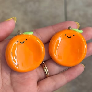 May include: Two orange-colored, ceramic dishes shaped like oranges, each with a smiling face, a green leaf, and a brown stem. The dishes are round and glossy, with a small indentation in the center.