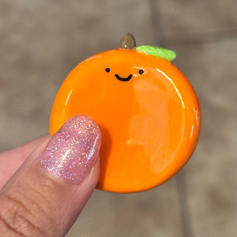 May include: A glossy orange-colored charm shaped like an orange with a smiling face. The charm has a small green leaf and a brown stem. The charm is being held by a person with a pink glitter nail.