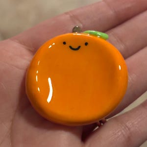 May include: A cheerful orange-colored pendant with a smiling face, a green leaf, and a silver bail. The pendant is round and glossy, resembling a cartoon orange. The design is simple and playful, suitable for a necklace or charm.