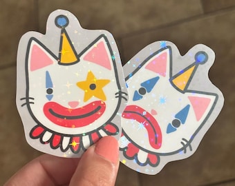May include: Two stickers featuring a cartoon cat with clown makeup. The cat has a white face, pink ears, and a red, white, and blue ruffled collar. The cat wears a yellow party hat and a star. The stickers have a holographic, glittery effect.
