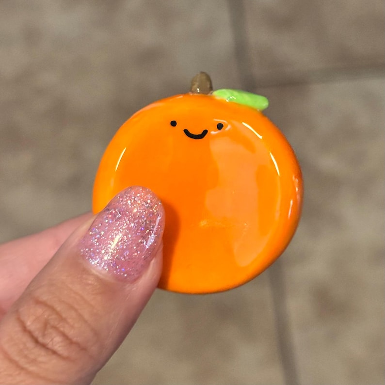 May include: A cheerful orange-colored pin with a smiling face, a small green leaf, and a brown stem. The pin is round and glossy, resembling a cartoon orange. The background is blurred.