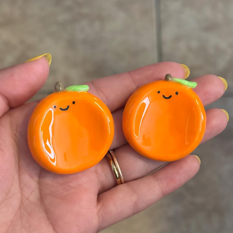 May include: Two orange-colored, ceramic dishes shaped like oranges, each with a smiling face, a green leaf, and a brown stem. The dishes are round and glossy, with a small indentation in the center.